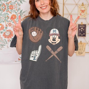 May include: A dark grey t-shirt with a baseball theme featuring a cartoon Mickey Mouse wearing a baseball cap with the letter "M" on it, a baseball glove with a baseball, a baseball bat and a hand with the number "1" on it.