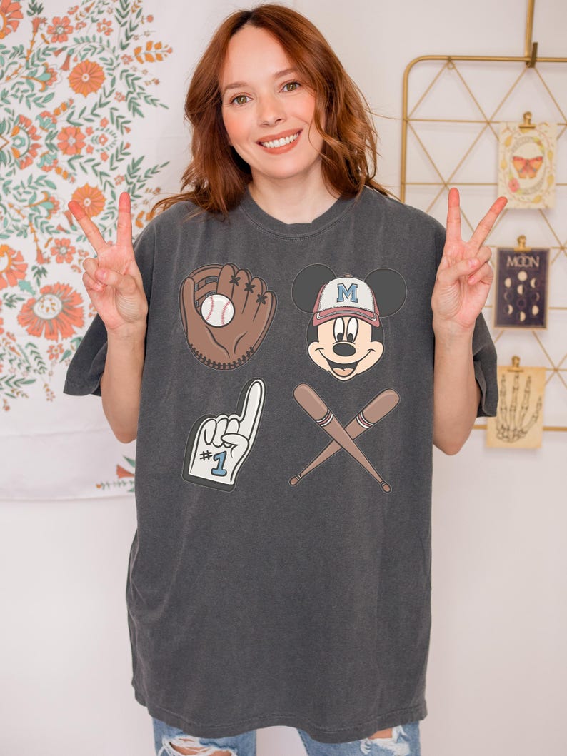 May include: A dark grey t-shirt with a baseball theme featuring a cartoon Mickey Mouse wearing a baseball cap with the letter "M" on it, a baseball glove with a baseball, a baseball bat and a hand with the number "1" on it.