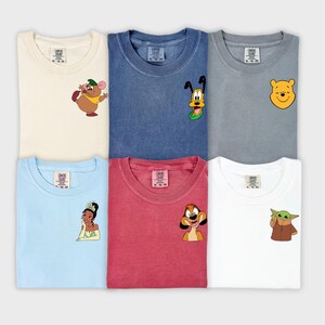 May include: Six colorful t-shirts with cartoon character designs. The shirts are cream, blue, gray, light blue, red, and white. Each shirt features a different character, including a mouse, Pluto, Winnie the Pooh, a princess, Timon, and Grogu.