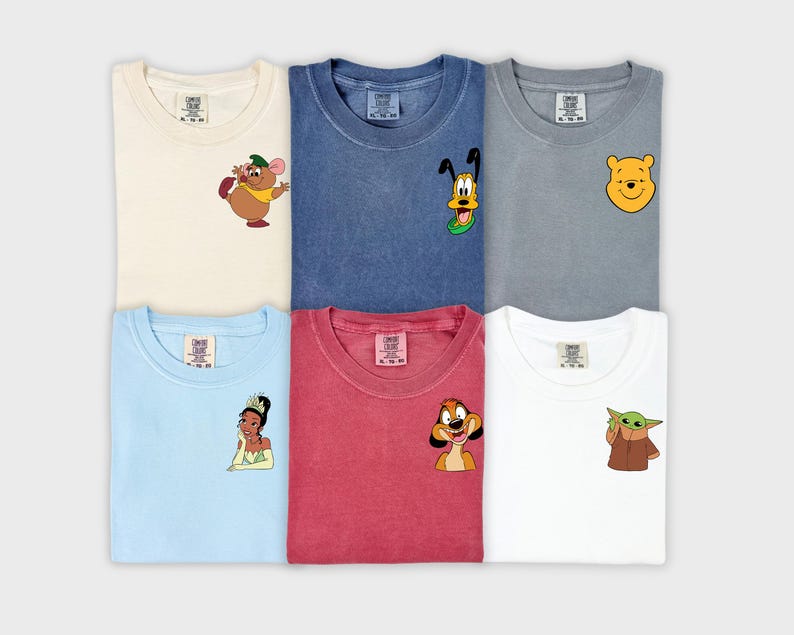 May include: Six colorful t-shirts with cartoon character designs. The shirts are cream, blue, gray, light blue, red, and white. Each shirt features a different character, including a mouse, Pluto, Winnie the Pooh, a princess, Timon, and Grogu.