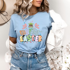 Puede incluir: Camiseta azul claro con el texto "my first EASTER" en letras coloridas. El gr&aacute;fico incluye un personaje de dibujos animados con orejas de conejo y un huevo de Pascua. La camiseta se usa con pantalones cortos de mezclilla azules.