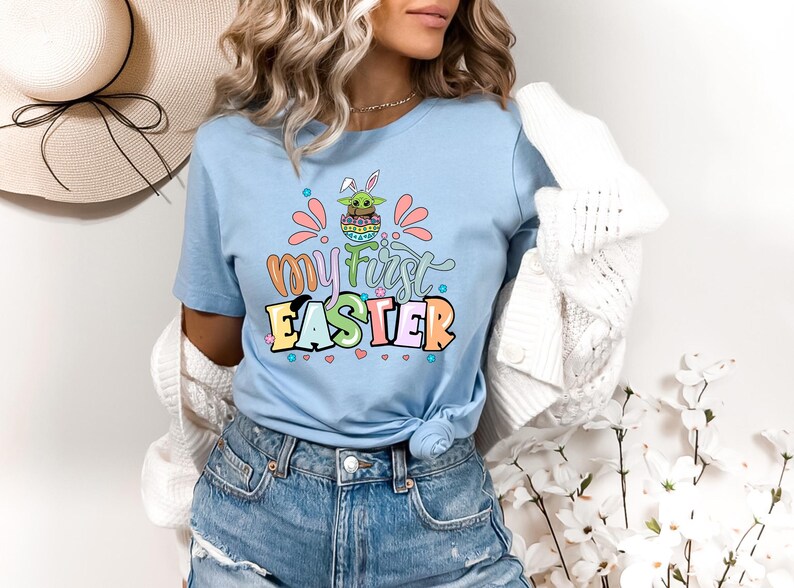 Puede incluir: Camiseta azul claro con el texto "my first EASTER" en letras coloridas. El gr&aacute;fico incluye un personaje de dibujos animados con orejas de conejo y un huevo de Pascua. La camiseta se usa con pantalones cortos de mezclilla azules.