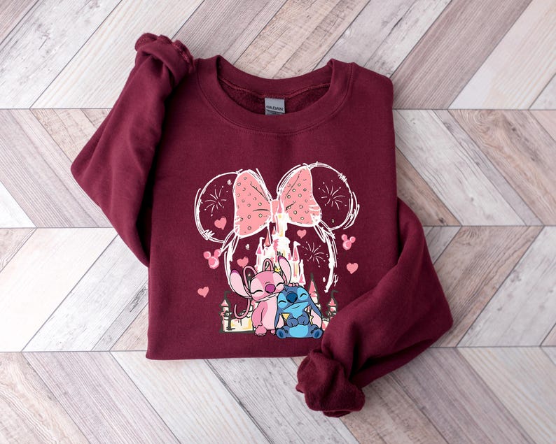May include: Burgundy sweatshirt with a Minnie Mouse-themed design, including a pink bow, castle, and cartoon characters. The graphic incorporates pink hearts and fireworks. A casual, comfortable garment.