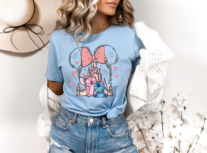 May include: Pale blue t-shirt with a cartoon graphic of a castle, pink bow, and characters. The design includes a pink and blue character, with heart and firework accents. The shirt is a short-sleeved crew neck.