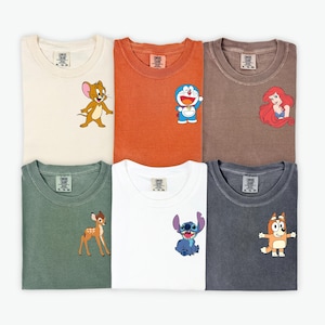 May include: Six folded t-shirts in various colors, each featuring a cartoon character on the front. Colors include cream, orange, brown, green, white, and charcoal. Characters include Jerry, Doraemon, Ariel, Bambi, Stitch, and Bluey.