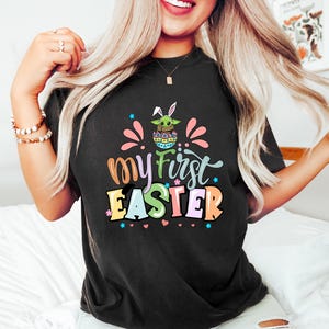 Puede incluir: Camiseta negra con el texto "My First Easter" en letras coloridas. El gr&aacute;fico incluye un personaje verde con aspecto de extraterrestre con orejas de conejo dentro de un huevo de Pascua. Acentos rosas y azules rodean el texto.