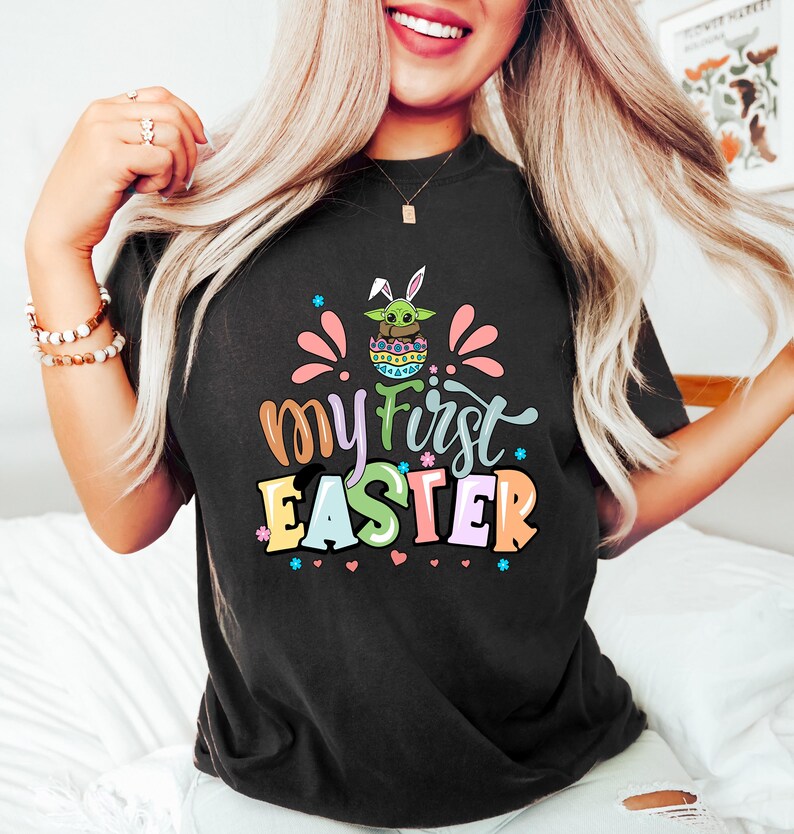 Puede incluir: Camiseta negra con el texto "My First Easter" en letras coloridas. El gr&aacute;fico incluye un personaje verde con aspecto de extraterrestre con orejas de conejo dentro de un huevo de Pascua. Acentos rosas y azules rodean el texto.