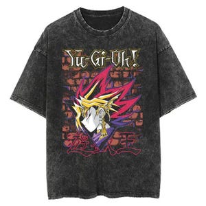 Pode incluir: T-shirt cinzenta escura com um gráfico da série de anime Yu-Gi-Oh!. O design inclui o texto "Yu-Gi-Oh!" e uma ilustração de personagem com detalhes em vermelho, roxo e dourado, sobre um fundo de parede de tijolos.