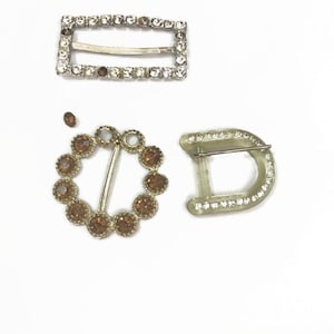 May include: Three vintage metal and plastic buckles with rhinestones. The buckles are in different shapes and sizes. One is a rectangle, one is a circle, and one is a D-shape.