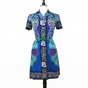 May include: A vintage blue dress with a floral and paisley print. The dress features short sleeves, a collared neckline, and a button-down front. A green belt cinches the waist. The dress is displayed on a mannequin.