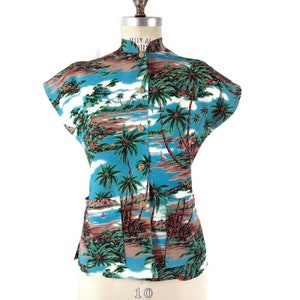 May include: A short-sleeved shirt with a tropical island print. The shirt has a mandarin collar and wooden buttons. The print features palm trees, blue skies, and ocean scenes. The shirt is displayed on a mannequin.