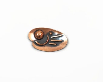 May include: A copper-toned brooch with a unique, abstract design. The brooch features a large, spherical element and a series of linear and curved accents. The brooch has a dark gray background and is set against a white background.