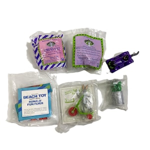 May include: A selection of McDonald's Happy Meal toys in their original packaging. The toys include a Bugs Bunny figure on a tricycle, a purple toy, and a small potted plant. Some packages have text.