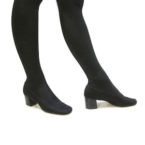 May include: Black over-the-knee boots with a low, block heel. The boots are made of a soft, stretchy material and have a rounded toe. The heels are approximately 2 inches high. The boots are shown against a white background.