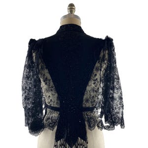 以下が含まれることがあります： 花柄のレース袖と、ビーズや刺繍が施された無地の黒いセンターパネルが特徴の黒いボレロジャケット。ハイカラーで、背面の裾にはビーズのフリンジがあしらわれています。