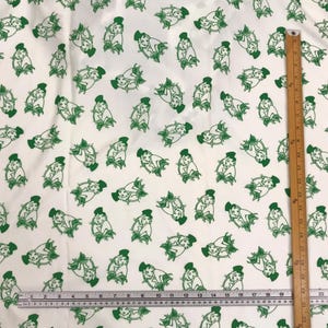 May include: White fabric featuring a repeating pattern of green figures. Each figure is a stylized illustration of a person in a hat and dress. A wooden ruler with cm markings is on the right side.