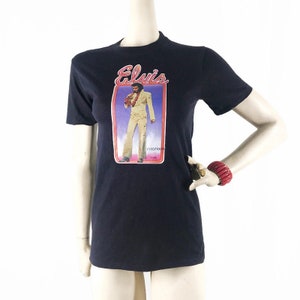 May include: Black t-shirt featuring a graphic of Elvis Presley in concert. The graphic includes the name "Elvis" in a stylized font and an image of Elvis in a white jumpsuit. The shirt is displayed on a mannequin.