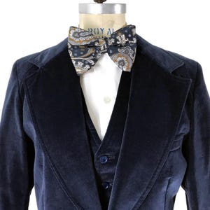 May include: A navy blue velvet blazer with a matching waistcoat and a paisley bow tie. The blazer has a shawl collar and a single button fastening.