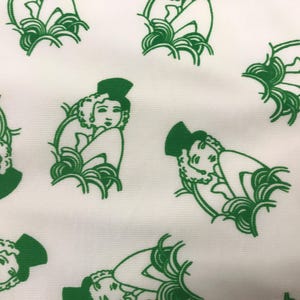 May include: White fabric featuring a repeating pattern of green Art Deco illustrations of women. Each figure is depicted with a hat and surrounded by stylized foliage. The design is a repeating pattern.
