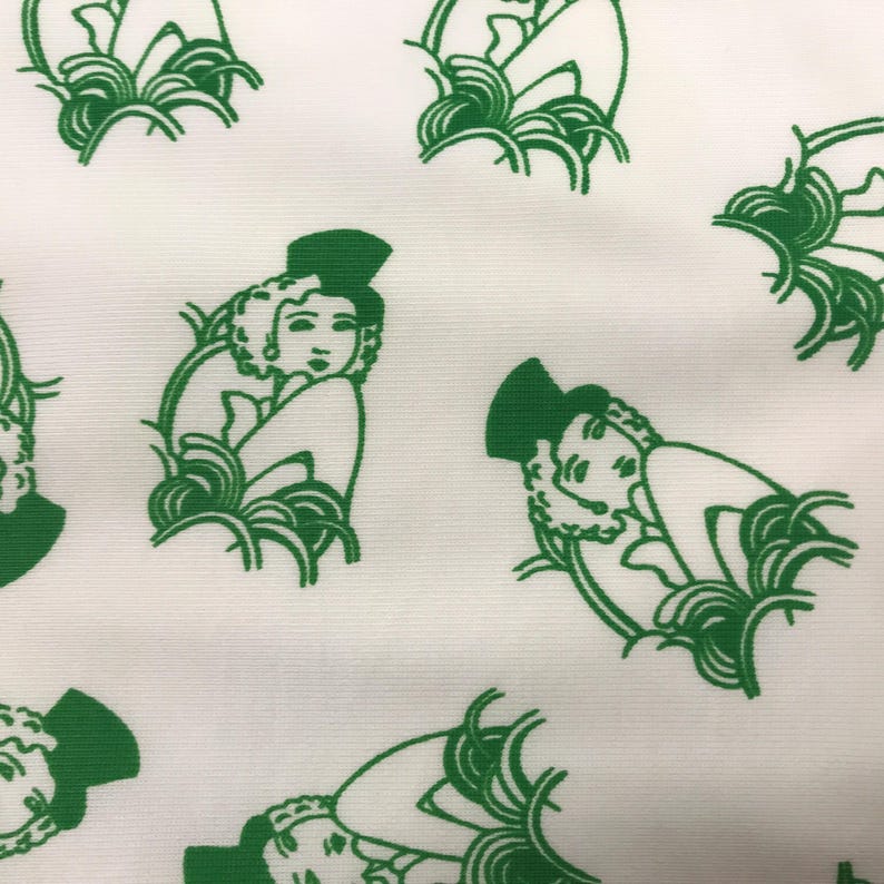 May include: White fabric featuring a repeating pattern of green Art Deco illustrations of women. Each figure is depicted with a hat and surrounded by stylized foliage. The design is a repeating pattern.