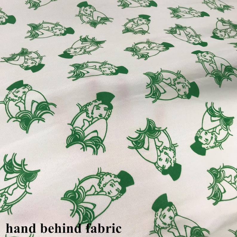 May include: White fabric featuring a repeating pattern of green illustrations. Each illustration shows a person in a top hat and a decorative design. The fabric has a soft, slightly textured appearance.