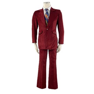 May include: A burgundy corduroy suit displayed on a mannequin. The suit includes a two-button jacket with pocket flaps and flared pants. A patterned tie and white dress shirt are visible.