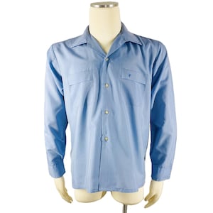 May include: Light blue long-sleeve shirt with a camp collar, two chest pockets, and a small embroidered emblem on the left pocket. The shirt has a button-down front and buttoned cuffs. The shirt is displayed on a mannequin.