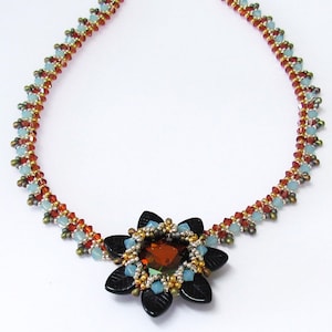 May include: A beaded necklace with a brown, blue, and gold color scheme. The necklace features a black flower-shaped pendant with a clear orange gemstone in the center.