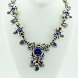 Op de afbeelding: Een kralenketting met een grote blauwe edelsteen hanger. De ketting is gemaakt met gouden, zilveren en paarse kralen. De ketting heet "Olivia" en is van Isabella Lam.