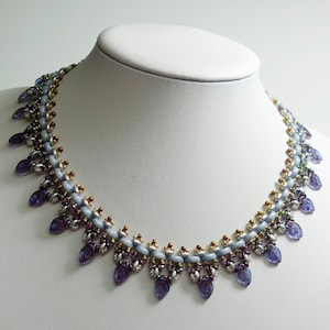 May include: A beaded necklace with a light blue base, adorned with purple leaf-shaped pendants. The necklace features gold, silver, and iridescent accents, creating a delicate and elegant design. The necklace is displayed on a white mannequin.