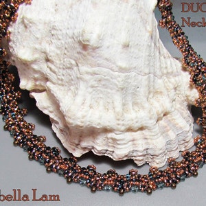 Op de afbeelding: Een koperen en blauwe kralenketting met de tekst "DUO Necklace" en "Isabella Lam" op een witte schelp achtergrond.