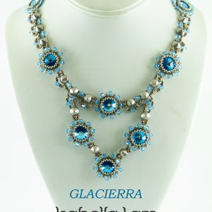 Puede incluir: Un collar de dos hileras con un esquema de color plateado y azul. El collar presenta un colgante central con un gran cristal azul rodeado de cristales más pequeños y cuentas plateadas. El collar se muestra en un busto de maniquí blanco. El texto "GLACIERRA Isabella Lam New Thinking with Endless Possibilities" se muestra debajo del collar.