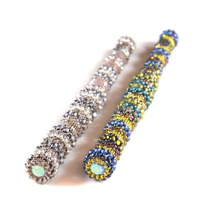 May include: Two beaded pens, one with silver and white beads and one with blue, green, and yellow beads. Both pens have a clear iridescent bead at the end.