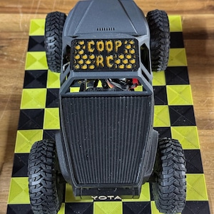 May include: A black and gray RC truck with large, textured tires. The truck has the text "COOP RC" on the front and "YOTA" on the front bumper. It sits on a yellow and black checkered surface.