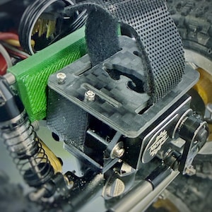 May include: Close-up of a black and gray RC car component. The component features a carbon fiber plate, a black strap, and a green section. The car's suspension and tire are visible in the background.