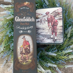 Può includere: Una scatola di metallo verde e dorata con le parole "Single Malt Glenfiddich Pure Malt Scotch Whisky" e un'immagine di un uomo in kilt. La scatola di metallo è da 750 ml e ha un'immagine di un golfista sul lato.