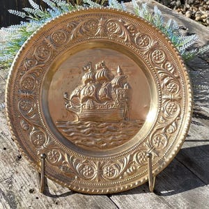 Könnte beinhalten: Eine goldfarbene Metallwandplatte mit einem geprägten Design eines Schiffes, das auf dem Meer segelt. Die Platte hat einen dekorativen Rand mit floralen und geschwungenen Mustern.