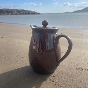 May include: A maroon ceramic teapot with a matching lid and handle rests on a sandy beach. The teapot has a dark brown rim and a light blue interior. The background shows the sea and a blue sky.