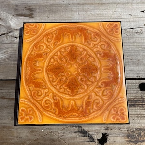 May include: Square ceramic tile with an intricate, embossed design in shades of orange and brown. The pattern features a central medallion surrounded by swirling motifs. The tile is set within a black frame, creating a decorative accent piece.