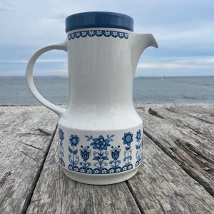 Op de afbeelding: Een witte keramische koffiepot met een donkerblauwe deksel en handvat. De pot heeft een blauw bloemmotief rond de basis en een decoratieve rand aan de bovenkant. De pot staat op een verweerd houten oppervlak.