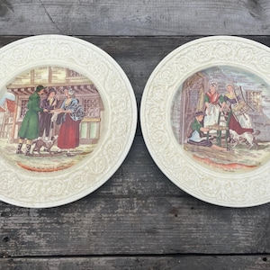 Może przedstawiać: Dwa białe ceramiczne talerze z ozdobnymi brzegami. Każdy talerz przedstawia kolorową ilustrację ludzi w historycznych strojach. Pierwszy talerz przedstawia mężczyznę i dwie kobiety na rynku. Drugi talerz przedstawia trzy kobiety pracujące w pokoju.