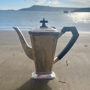Peut inclure: Une théière vintage argentée avec une poignée noire et un bouton en forme de croix sur le couvercle. La théière est rectangulaire avec un bec et est posée sur une plage de sable avec l'océan en arrière-plan.
