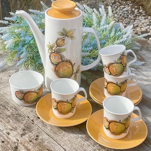 Pode incluir: Um conjunto de café vintage em cerâmica branca com uma cafeteira alta, seis xícaras e pires. O conjunto apresenta um design de maçã amarela e verde. A cafeteira tem uma tampa e uma alça amarelas. As xícaras e os pires são amarelos.