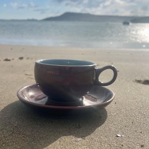 May include: A brown ceramic teacup and saucer set with a dark blue interior, resting on a sandy beach. The cup has a rounded shape with a handle. The background features the ocean and a distant landmass under a cloudy sky.