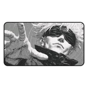 May include: A rectangular desk mat featuring a black and white manga-style illustration of a character with spiky hair and a blindfold over their eyes. The character is reaching forward with an open hand against a background of circular lines.