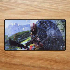 May include: A rectangular gaming mousepad depicting a warrior with a red stripe on his head, wielding a black blade and a large, dark shield. The background shows a mountainous landscape with a blurred effect.