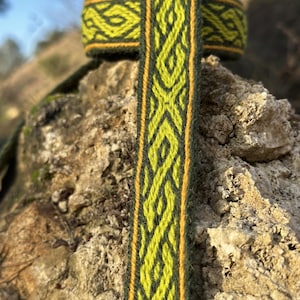 May include: Close-up of a woven green and yellow patterned ribbon. The ribbon has a dark green background with a repeating geometric design in yellow and a thin yellow border. The ribbon is resting on a textured rock surface.