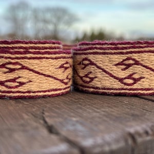 May include: Close-up of two rolled woven bands. The bands feature a tan background with a repeating burgundy pattern. The edges are also burgundy. The bands are resting on a weathered wooden surface.