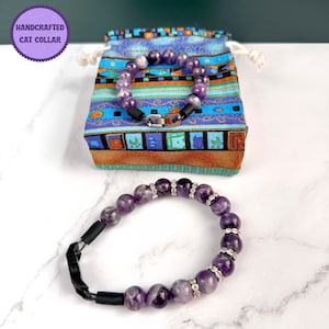 May include: A purple amethyst gemstone cat collar with a silver clasp and a black adjustable strap. The collar is in a small, colorful drawstring bag with the text "HANDCRAFTED CAT COLLAR".