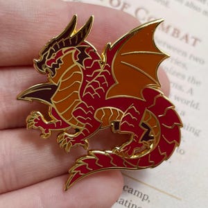 K&ouml;nnte beinhalten: Ein detaillierter Emaille-Pin mit einem roten und goldenen Drachen mit ausgebreiteten Fl&uuml;geln. Der Drache hat aufwendige Schuppen und einen goldenen Umriss. Der Pin ist ein Accessoire mit Fantasy-Thema, perfekt f&uuml;r Jacken, Taschen oder Sammlungen.